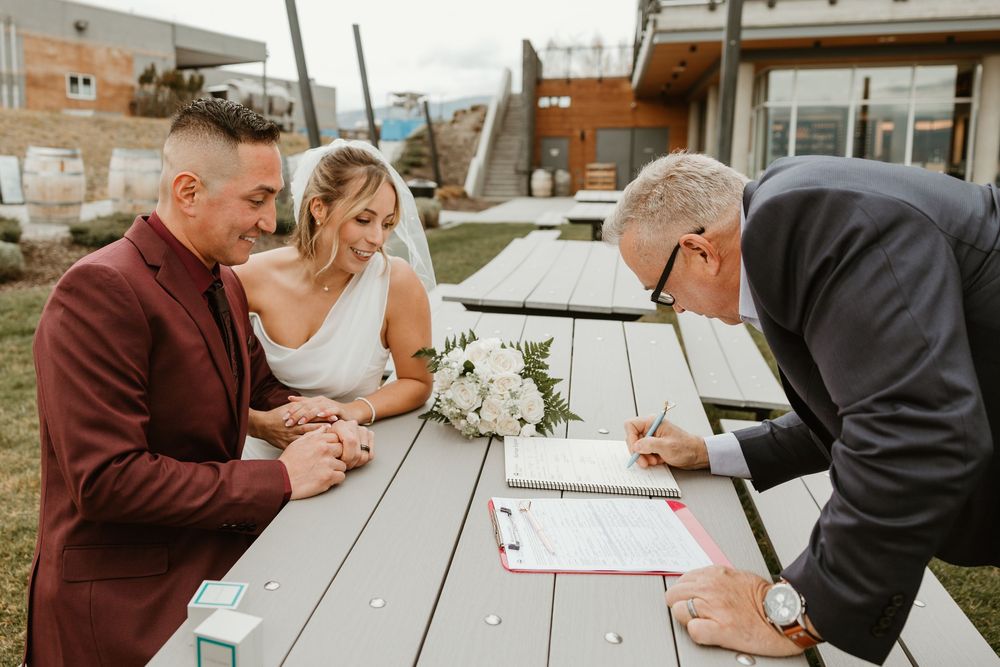 bride and groom smile as their witness signs their wedding paperwork at kelowna winery elopement