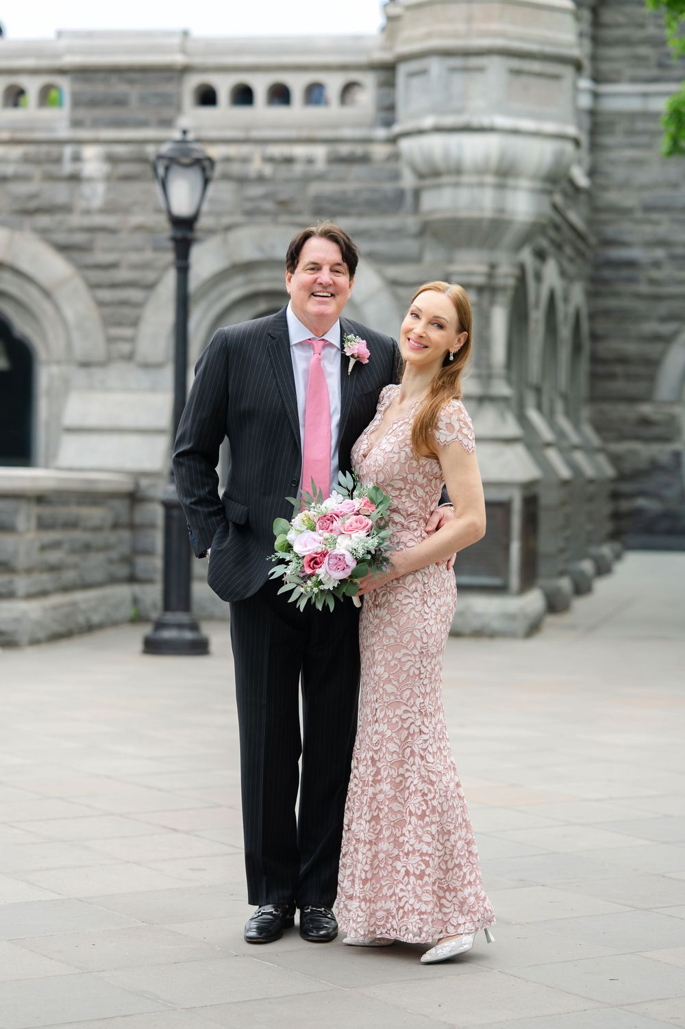 Couple in formal wedding attire posing outdoors near a stone building, woman holding pink floral bouquet.