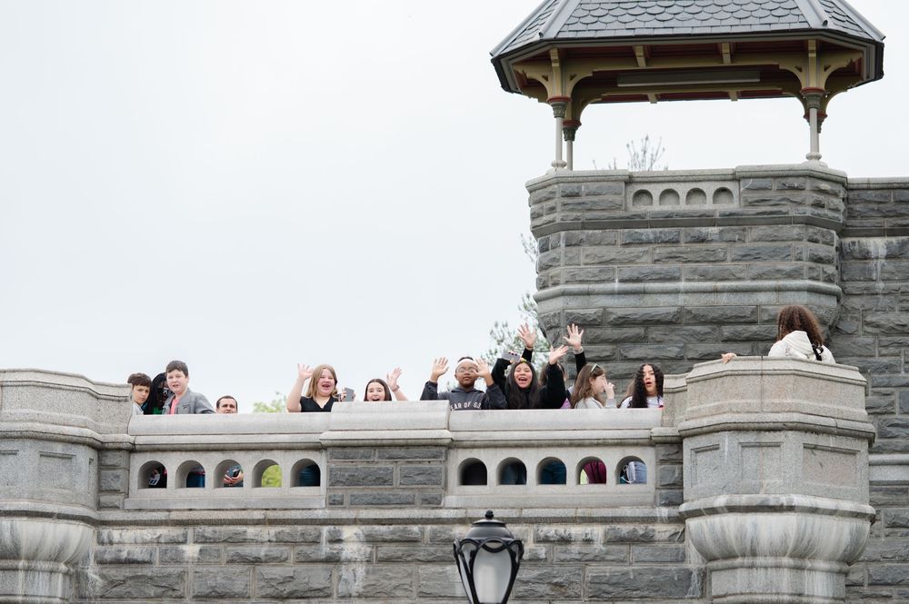 Group of tourists waving from the battlements of a historic stone castle on an overcast day.