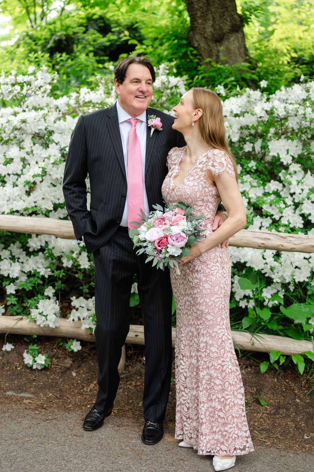 Couple in pink and black pinstripe suit poses with bouquet before white flowering bushes in park.
