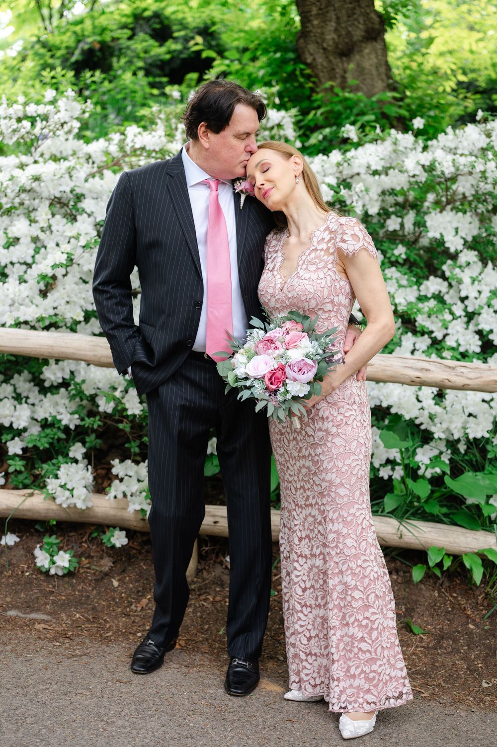 Couple in pink lace gown and pinstripe suit share tender moment before white flowering bushes.