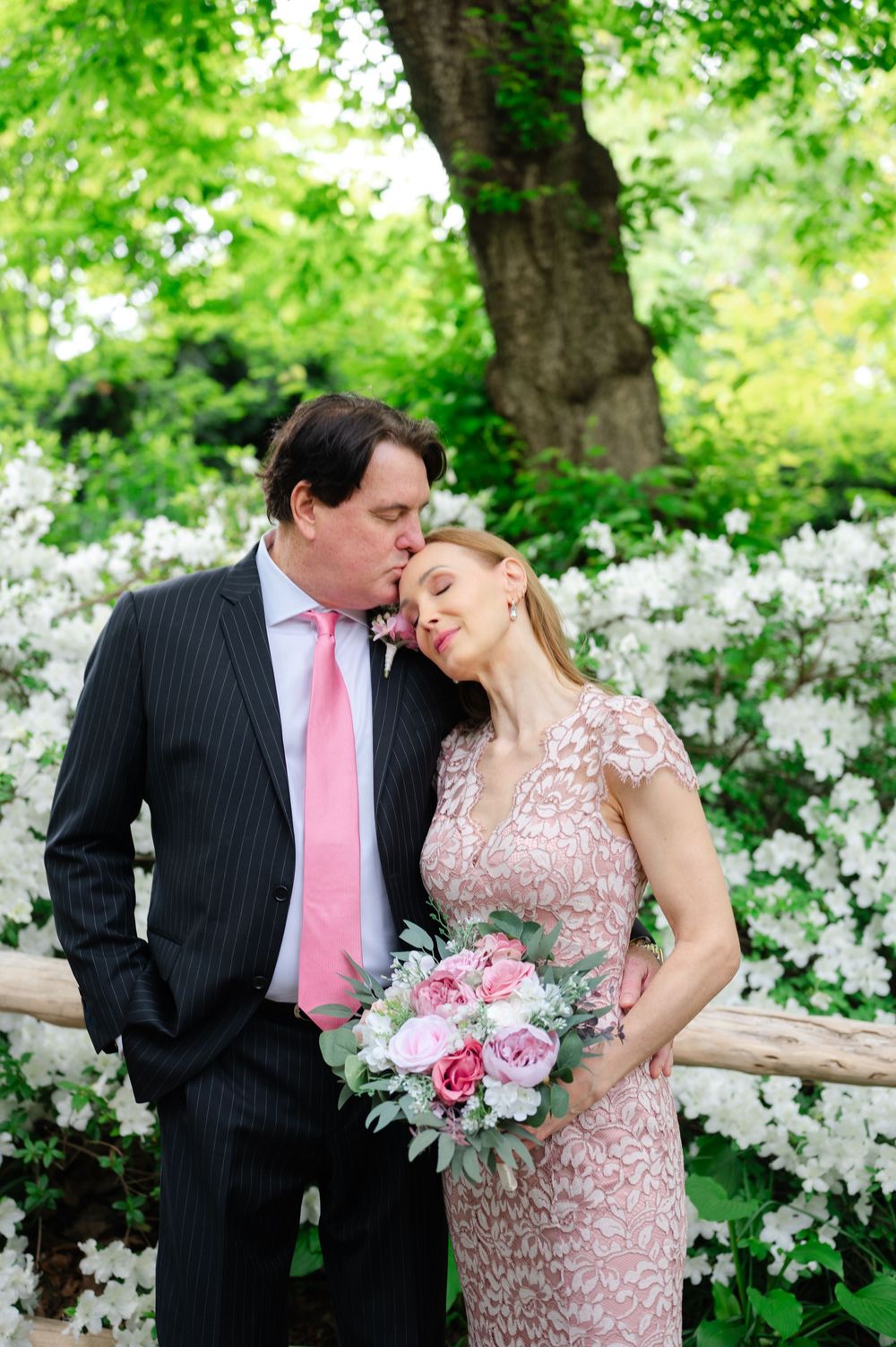 Couple sharing a tender kiss among white blooming flowers, groom in pinstripe suit with pink tie, bride in lace dress holding bouquet.