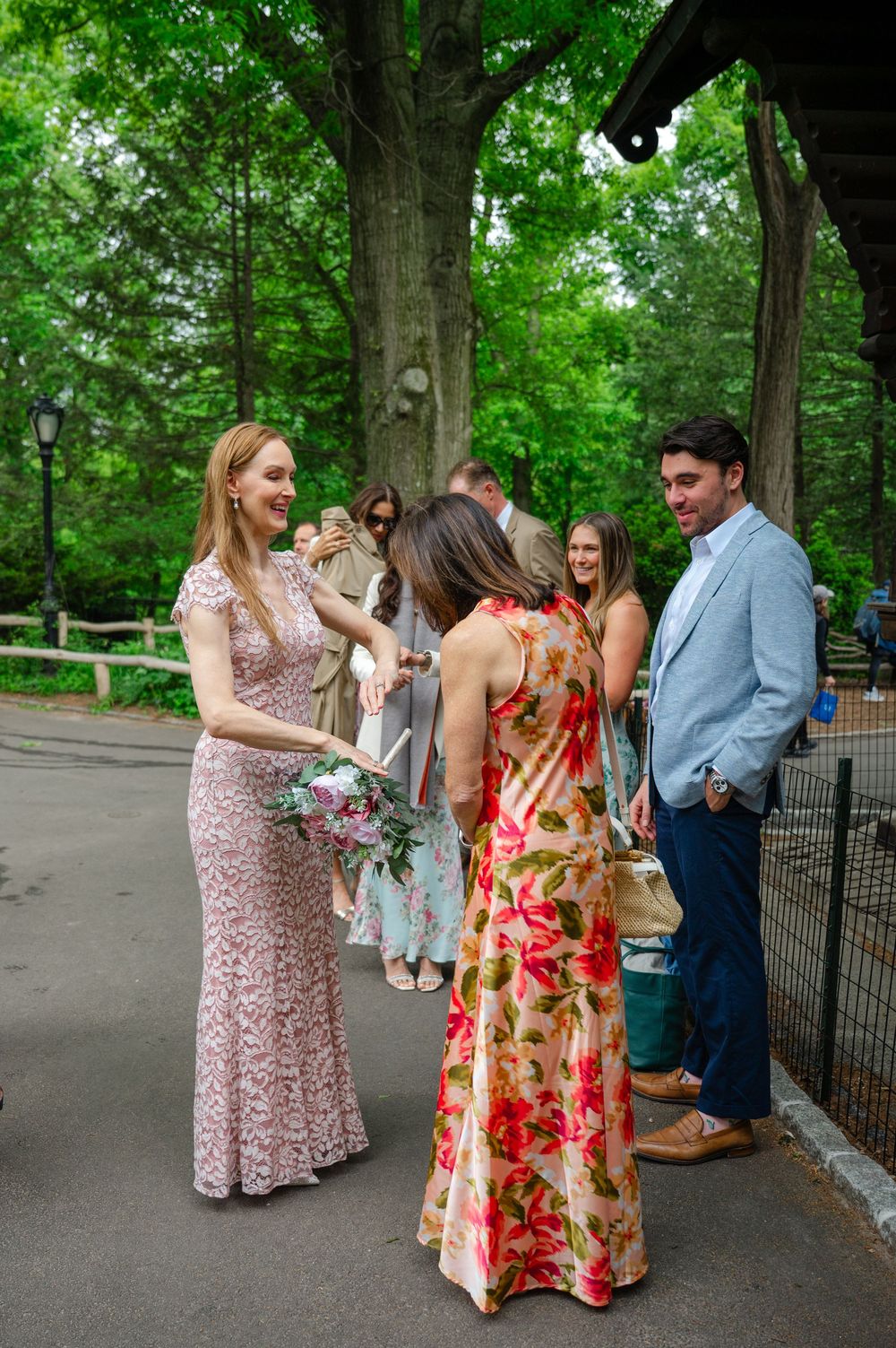 Bridesmaids in floral and lace dresses chat with guests at an outdoor wedding in a lush green park.