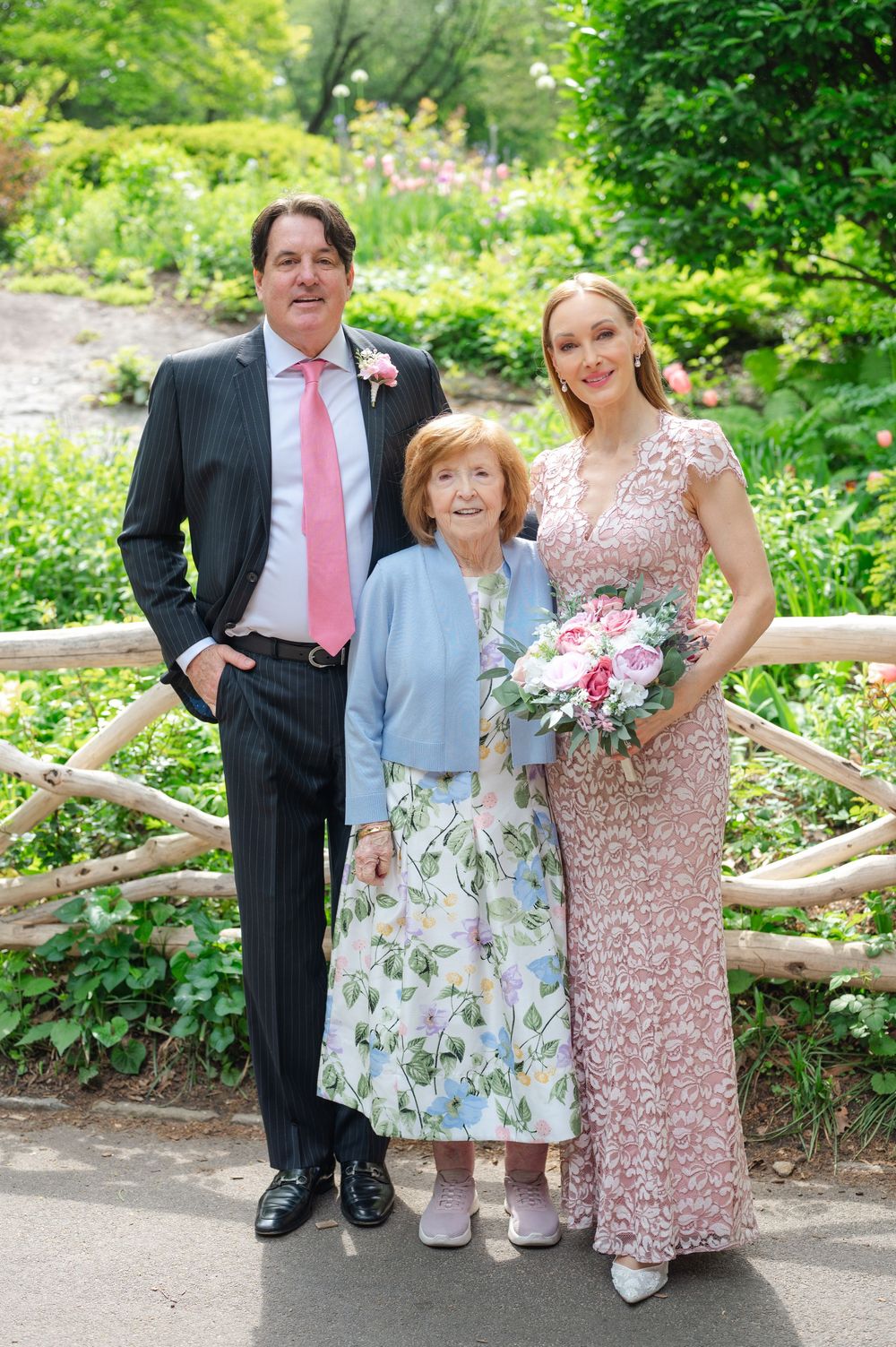 Wedding couple poses with elderly woman in floral dress in lush garden setting.