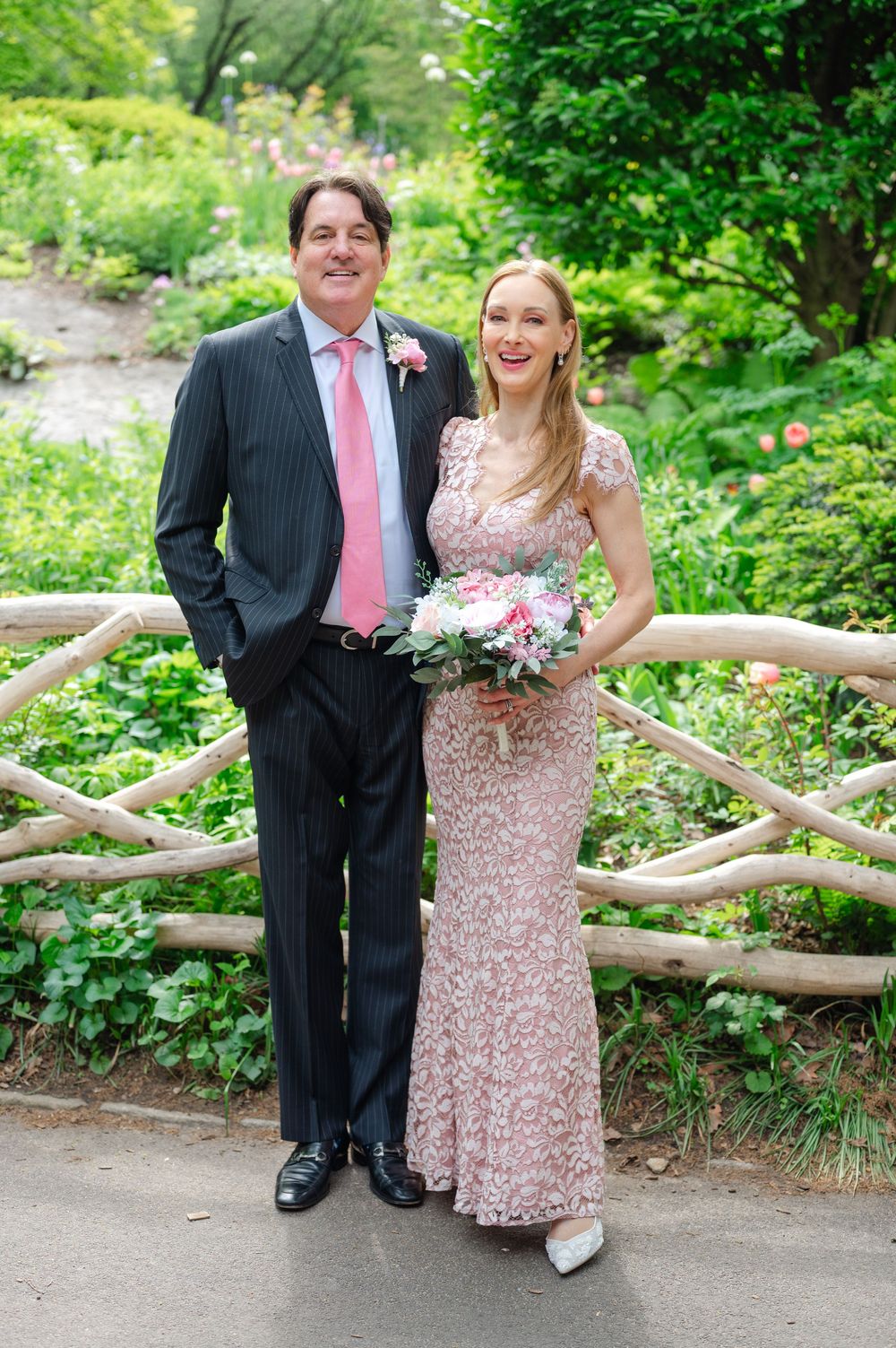 Smiling bride in pink lace dress and groom in pinstripe suit pose in garden.