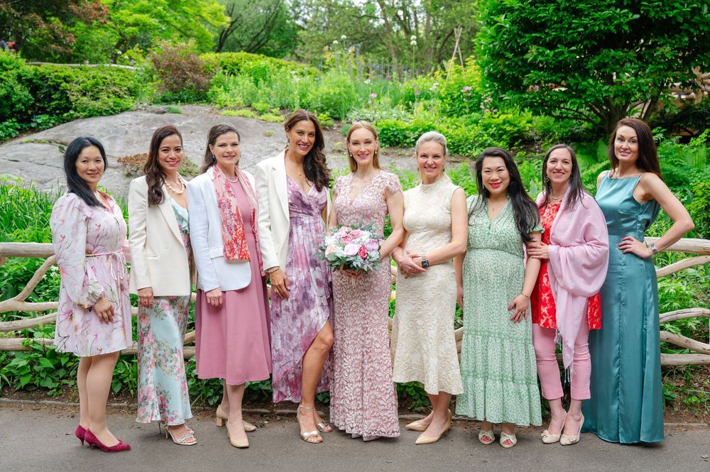 Group of eight women in elegant dresses posing together outdoors at a garden wedding celebration.