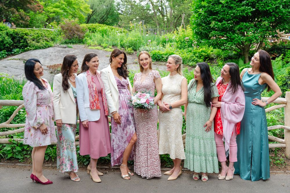 Eight women in elegant pastel dresses pose together laughing in a lush garden setting at a wedding celebration.