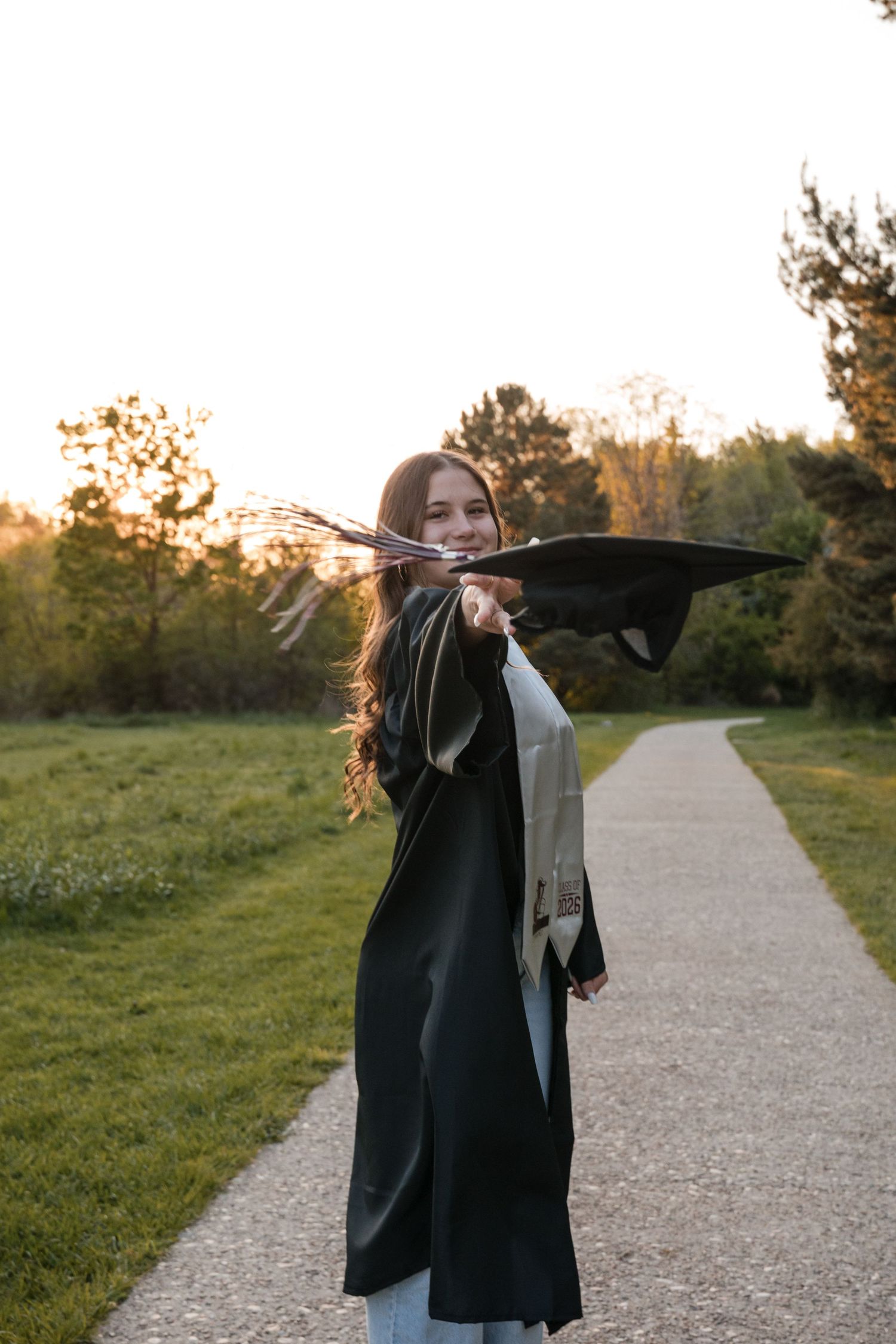 Young woman smiling outdoors at sunset, wearing jeans and open graduation gown on a tree-lined path.