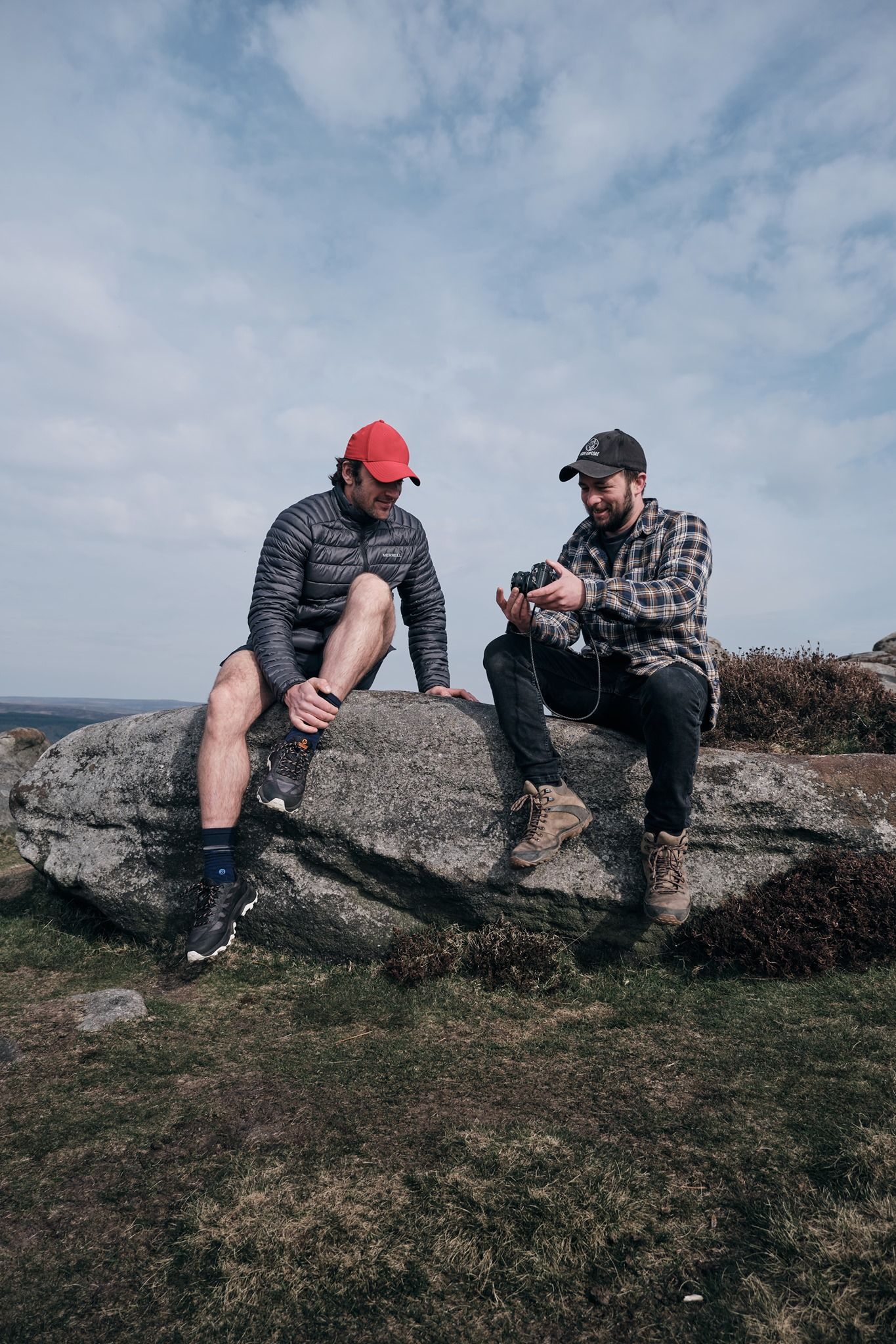 Two men sitting on rocks outdoors, one holding a camera, both wearing caps, with a cloudy sky backdrop.