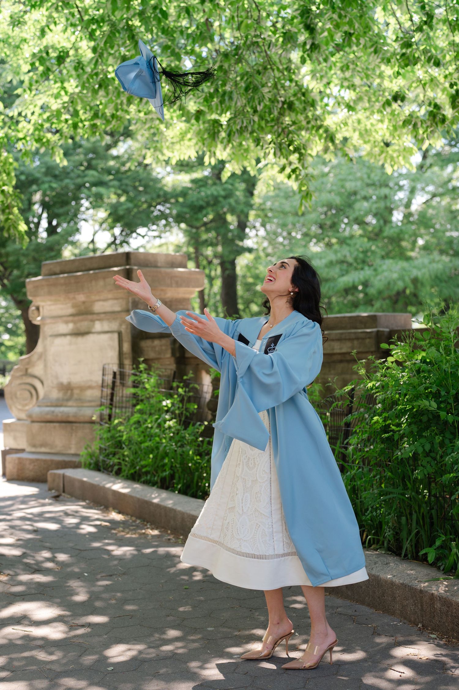 Columbia University Graduate in blue coat tossing graduation cap in lush green park, celebrating academic achievement in Riverside Park.