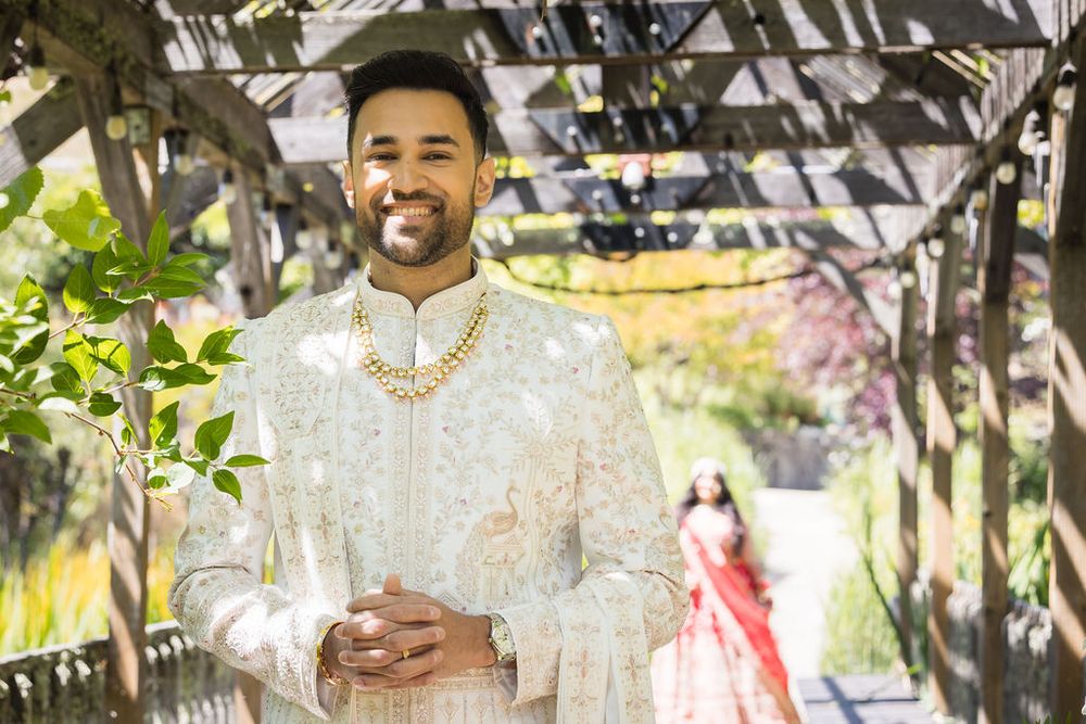 Groom in white embroidered sherwani with gold necklace smiles under pergola, bride in red lehenga visible in background.