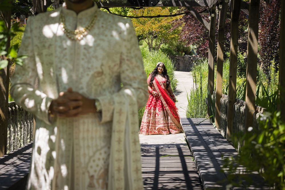 Indian bride in red lehenga stands on a garden path as groom in white sherwani waits in foreground.