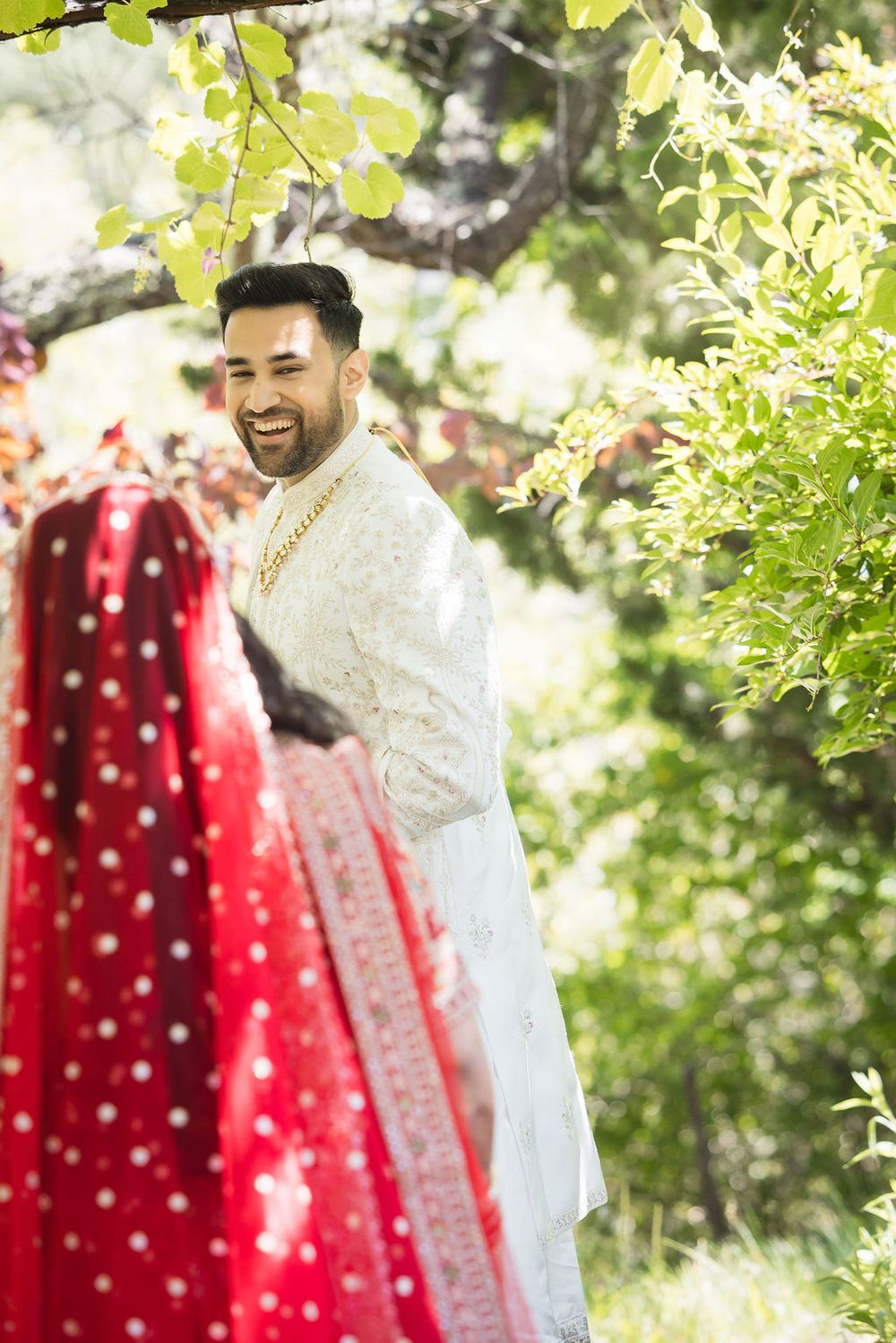 Smiling groom in white sherwani stands outdoors beside bride in red lehenga during Indian wedding ceremony.