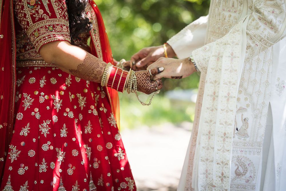 Indian bride in red lehenga with gold embroidery holds hands with groom during traditional wedding ceremony.