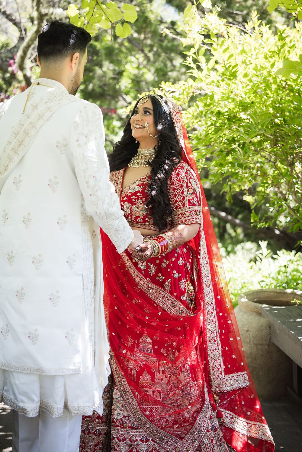 Indian bride in red lehenga with gold embroidery smiles at groom in white sherwani during outdoor wedding ceremony.