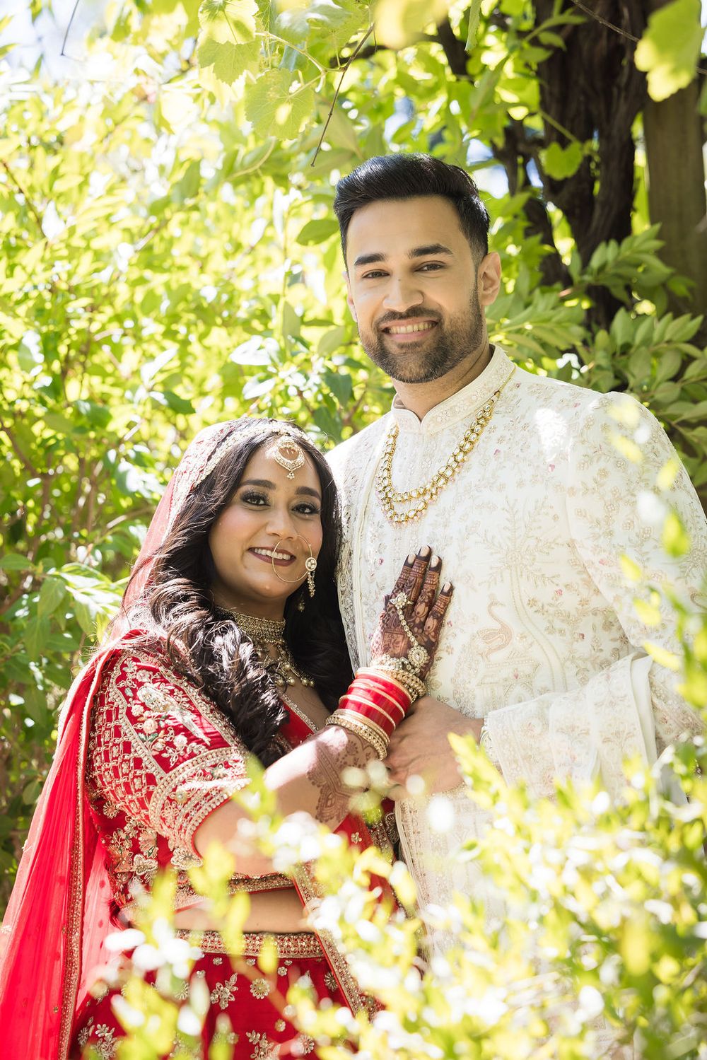 Indian bride in red lehenga and groom in white sherwani posing together outdoors surrounded by lush green foliage.