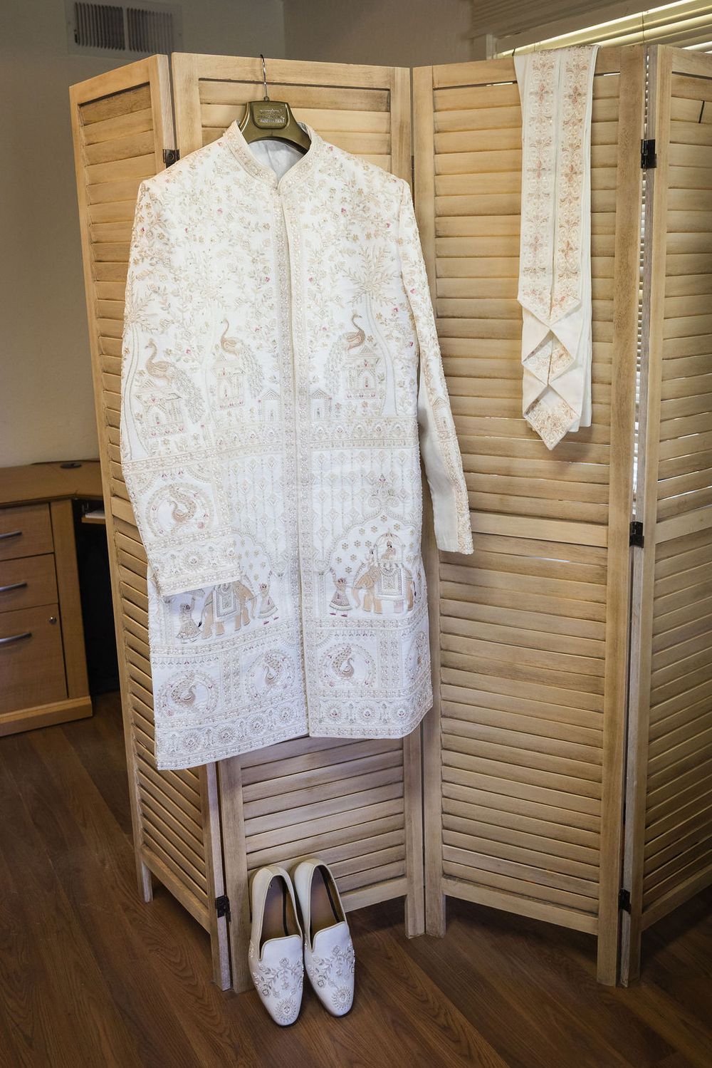 Elegant white embroidered sherwani with intricate gold detailing displayed on wooden room divider with matching shoes.