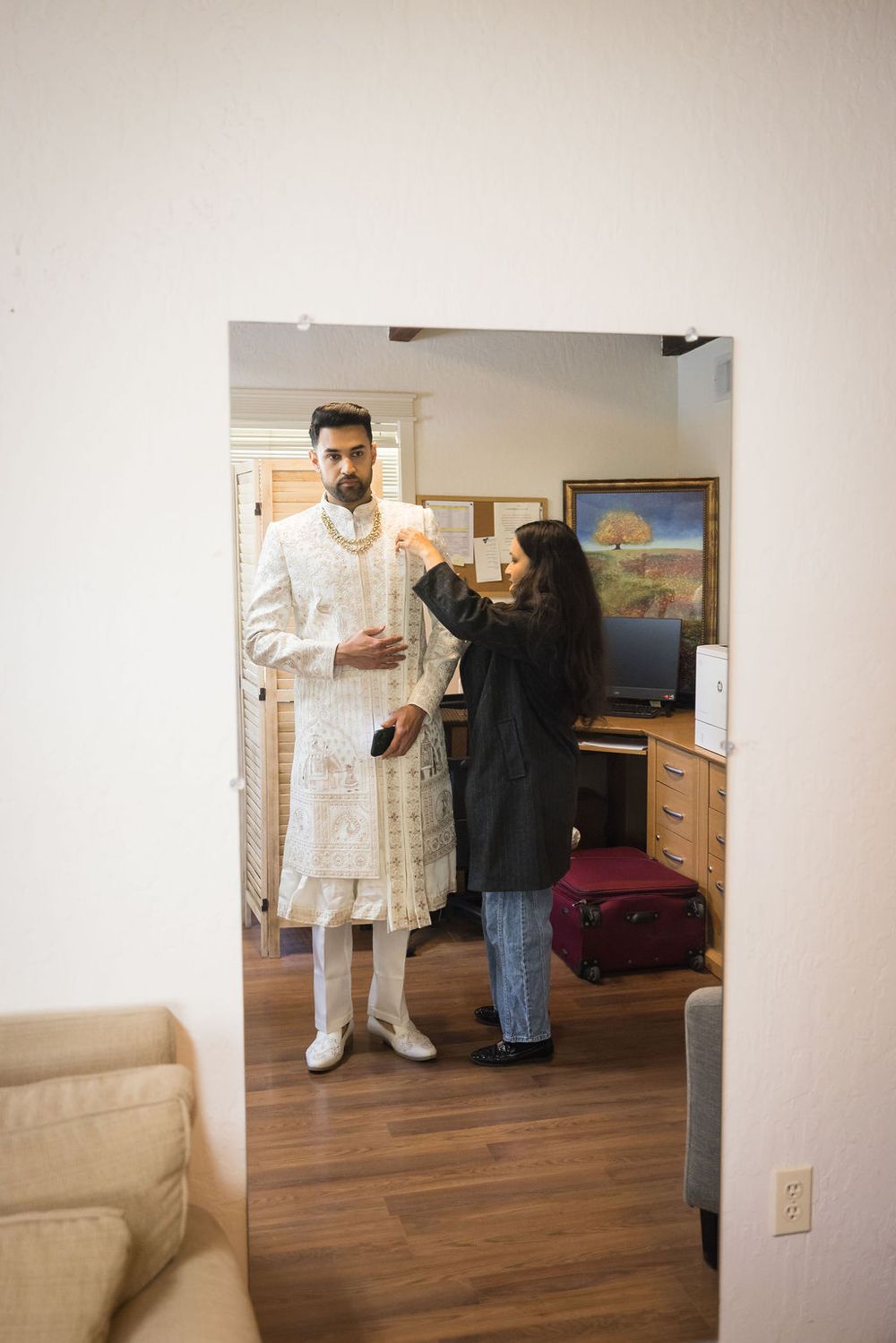 A woman helps a groom adjust his traditional white Indian sherwani wedding attire indoors.
