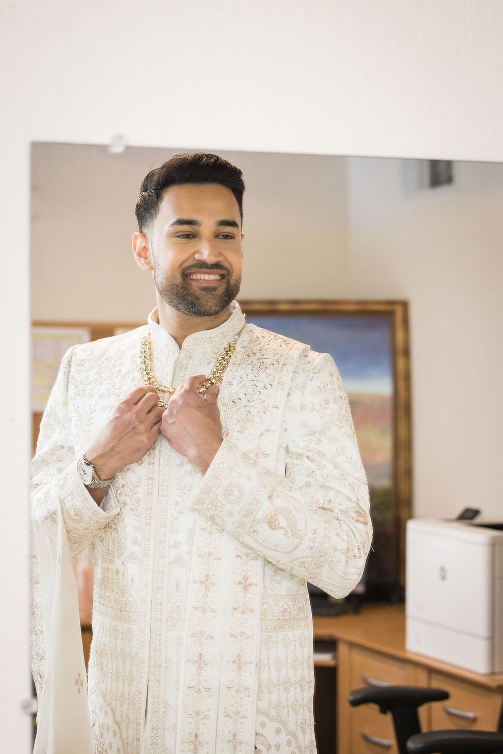 Smiling South Asian groom in ornate white and gold sherwani adjusting his collar before wedding ceremony.