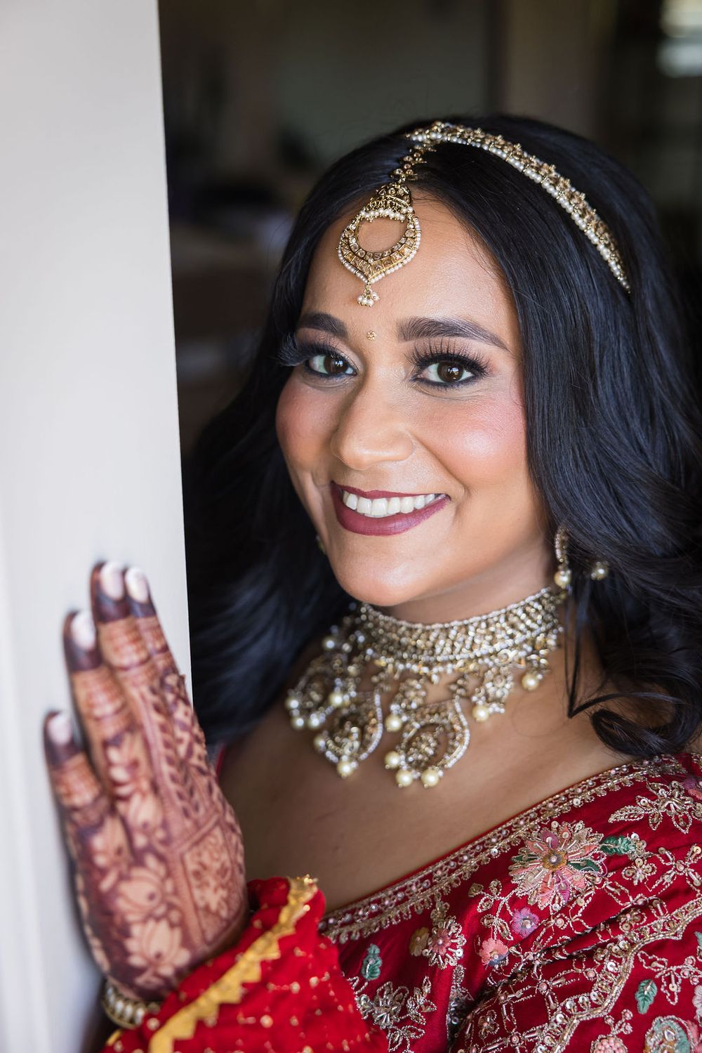 Beautiful Indian bride in red lehenga smiling with gold maang tikka, choker necklace, and intricate mehndi hands.