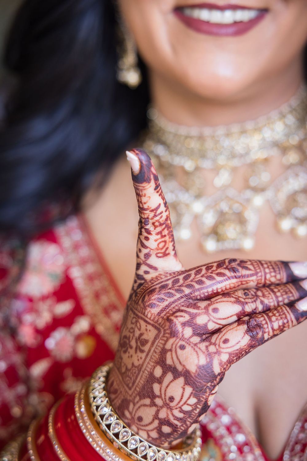 Indian bride showcasing intricate henna mehndi designs on her hands, wearing gold jewelry and red bridal outfit.