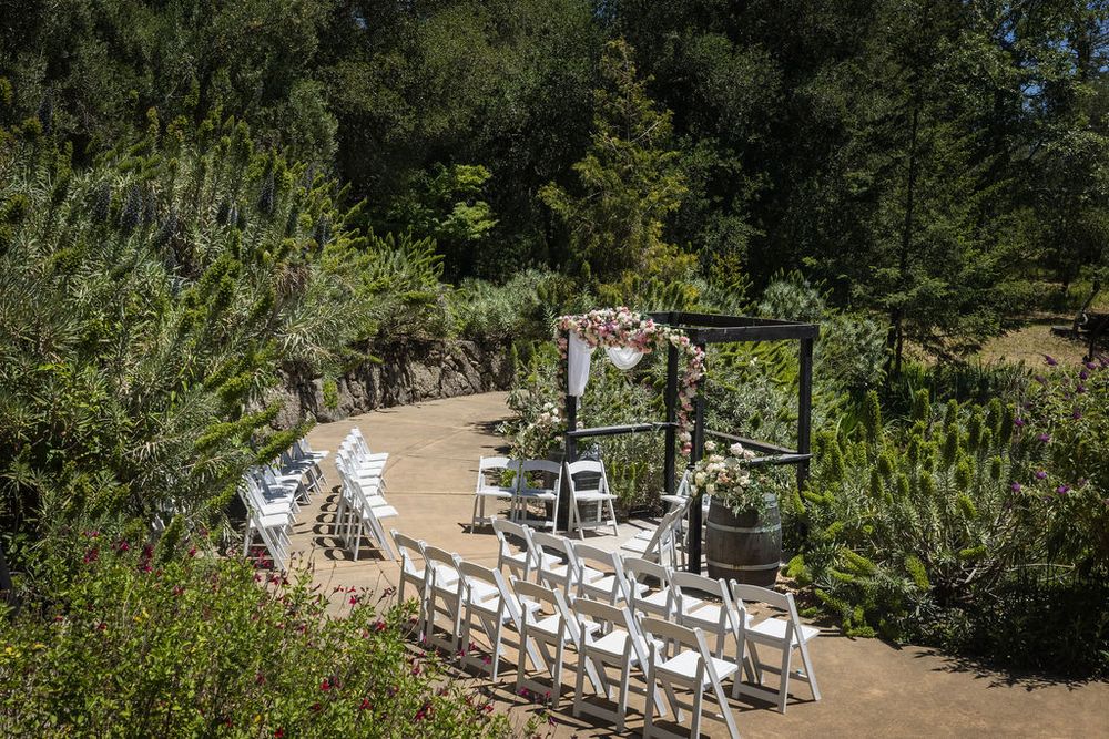Outdoor Indian wedding ceremony setup with white chairs and floral mandap arch surrounded by lush gardens.
