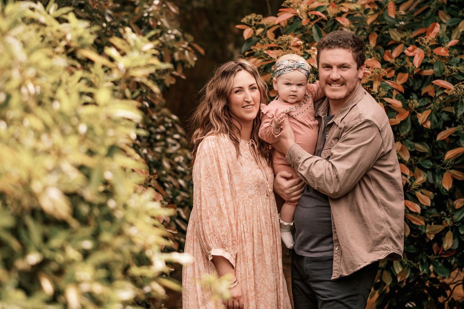 A smiling family of three poses outdoors, with a woman in a pink dress, a man in a tan shirt, and a baby girl.