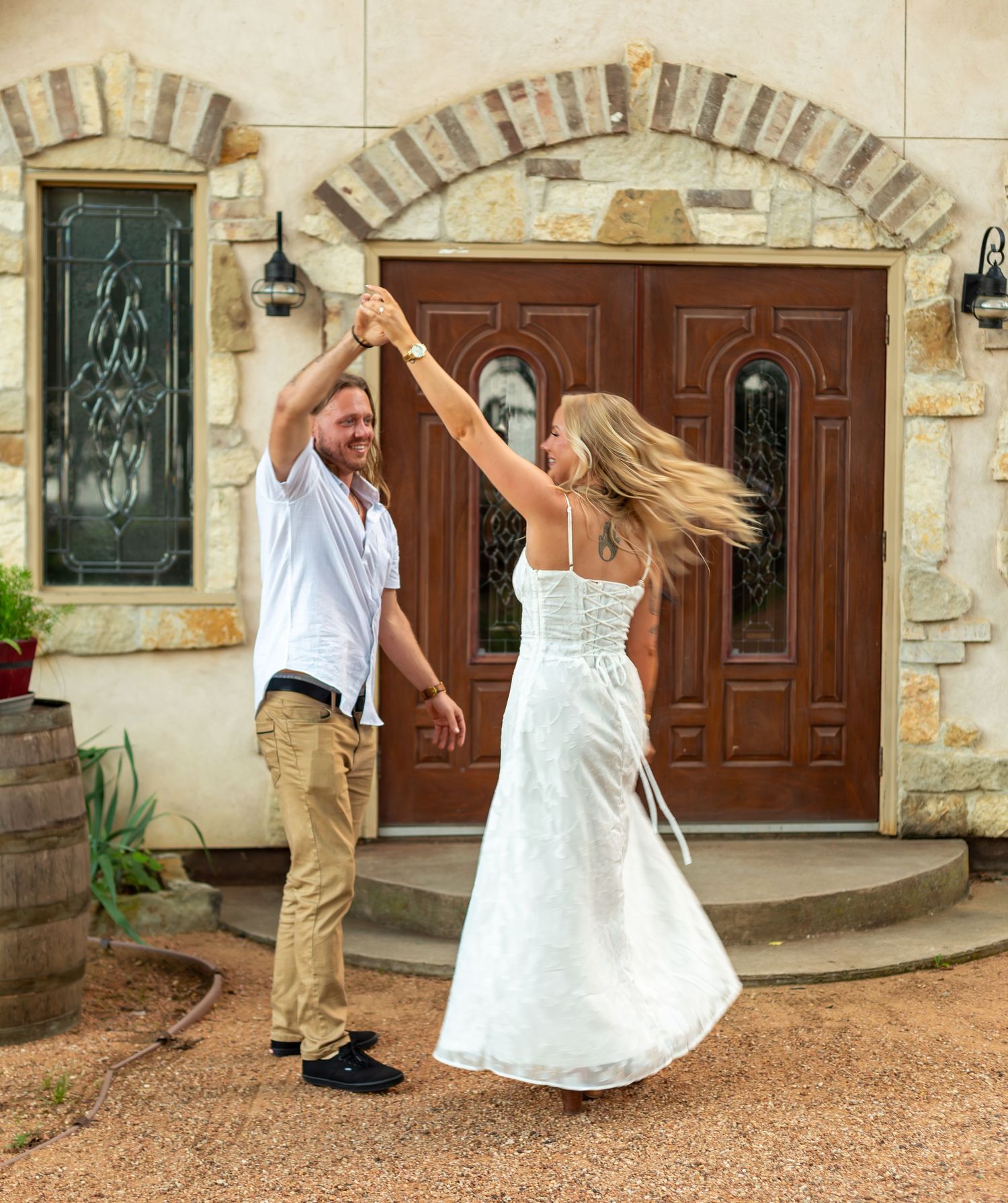 A couple dances joyfully outside elegant stone doors, woman in white dress twirling, man in white shirt and khaki pants.