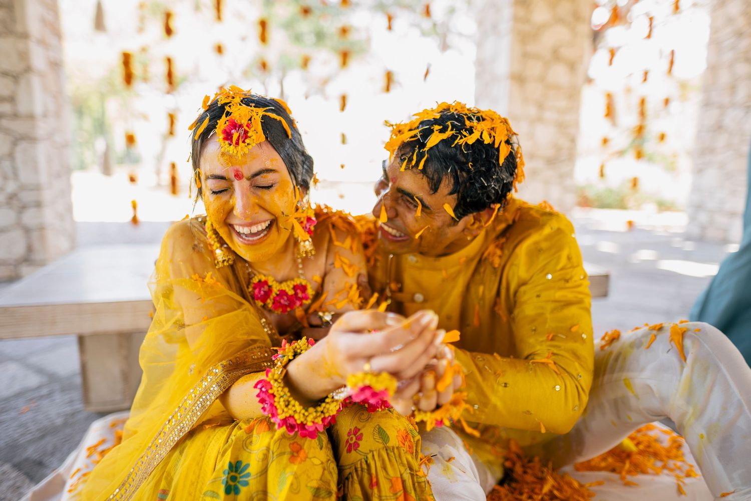 Joyful couple in yellow outfits celebrating Haldi ceremony with turmeric and flower petals.