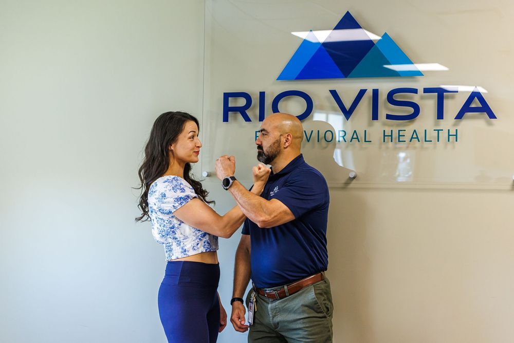 Two professionals interact warmly in front of Rio Vista Behavioral Health office sign.