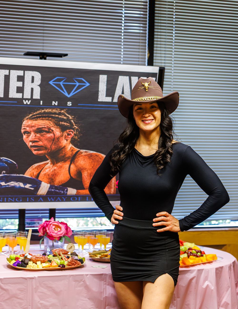 Woman in black dress and brown cowboy hat posing at event table with food and boxing promotional banner.