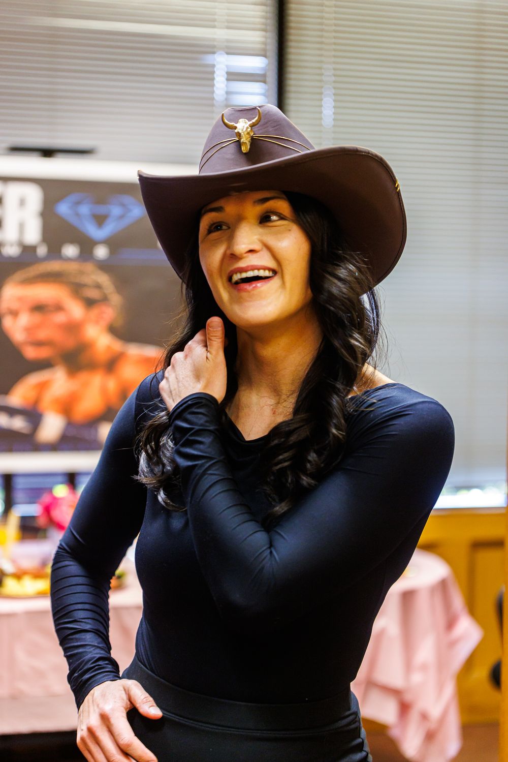 Smiling woman in black outfit and brown cowboy hat with bull emblem at indoor boxing event.