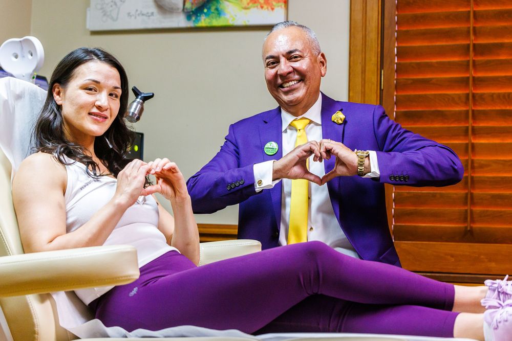 A woman and man in purple outfits make heart shapes with their hands while sitting together smiling.