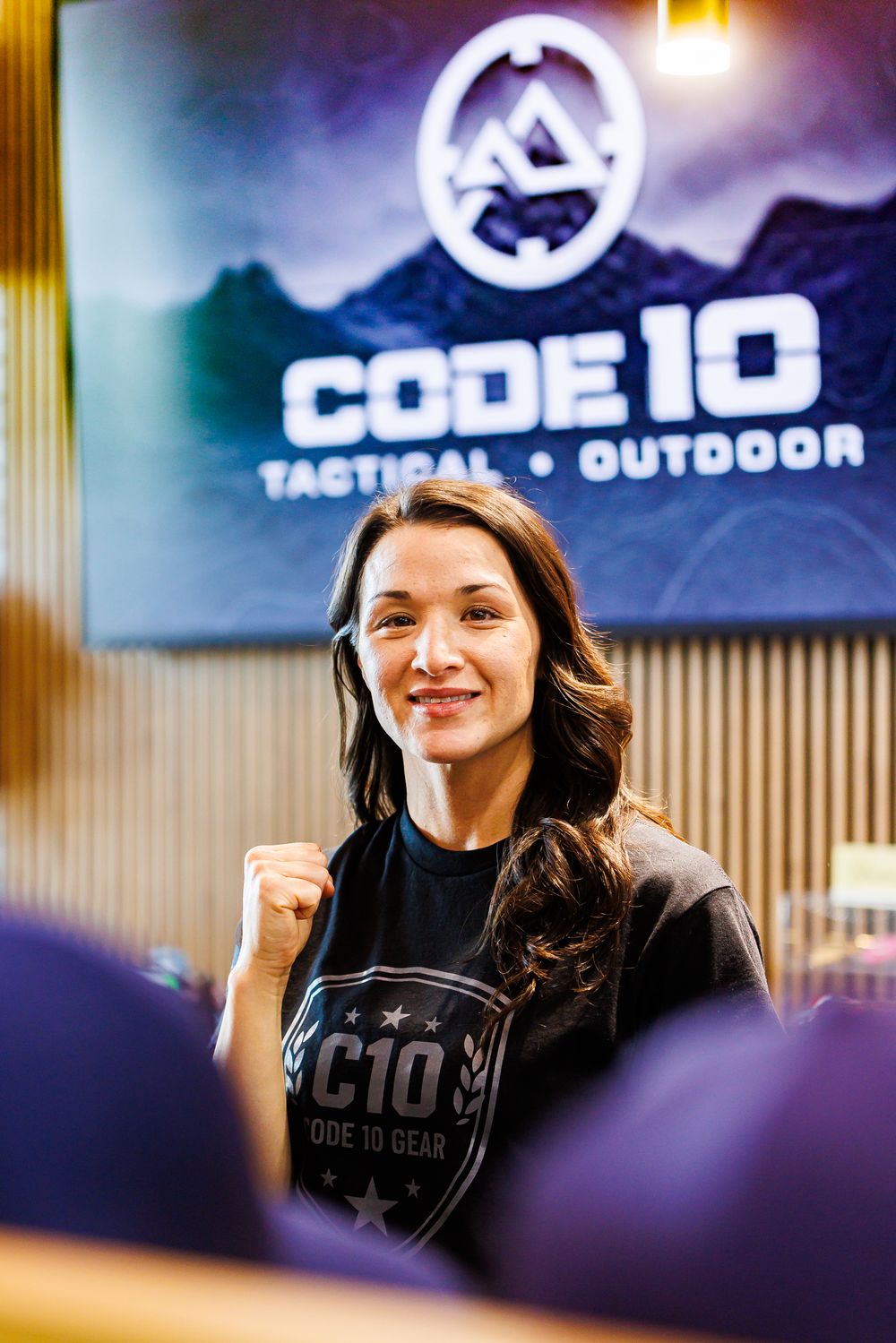 Athletic woman in Code 10 Tactical Outdoor t-shirt posing with raised fist at a retail store event.