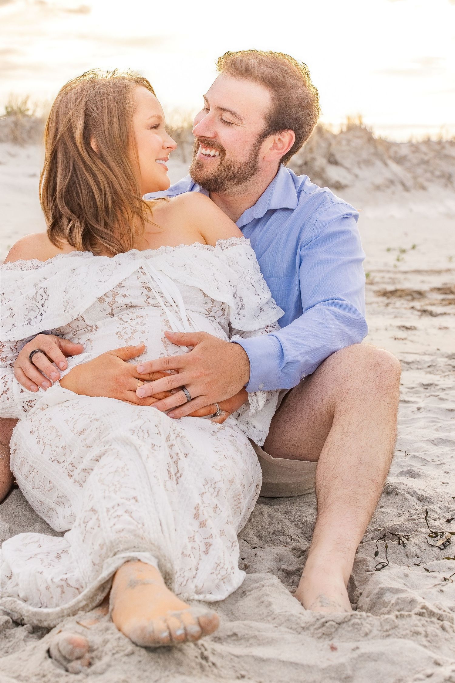 Couple sitting on beach sand, smiling at each other, woman in white lace dress, man in blue shirt, golden sunset light.