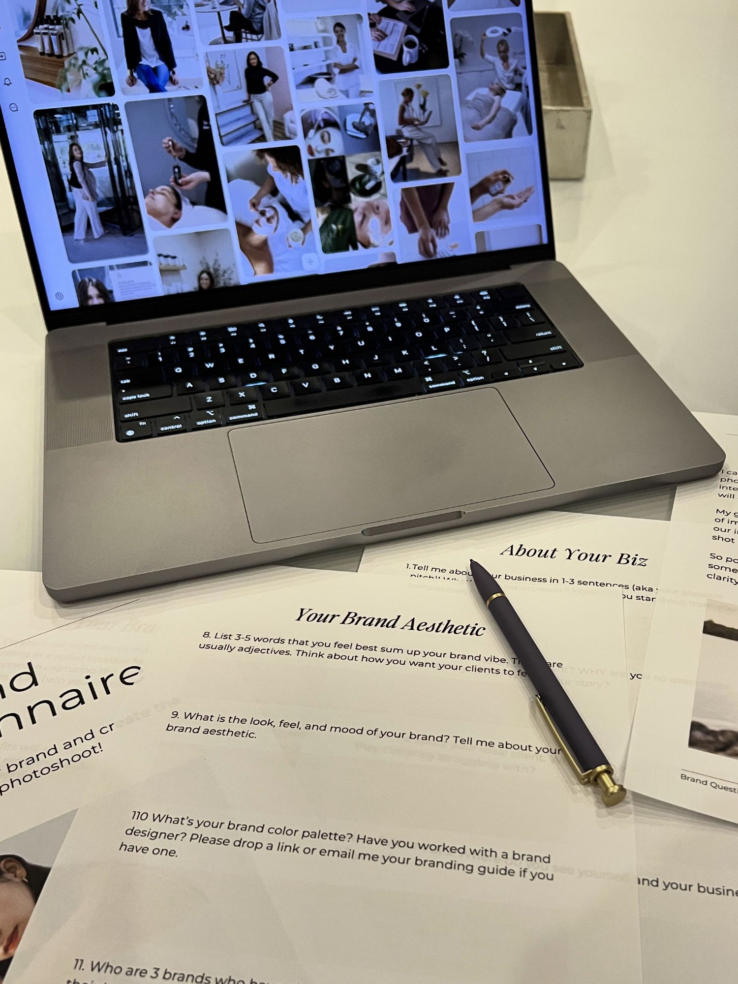 MacBook laptop displaying mood board photos beside brand questionnaire worksheets and a pen on a white desk.
