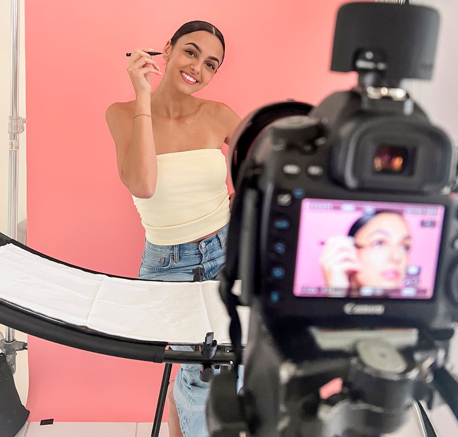 Woman applying makeup in front of a Canon camera, filming a beauty tutorial against a pink background.