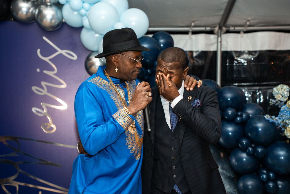 Two men in formal attire share an emotional moment at a festive event with blue and silver balloon decorations.