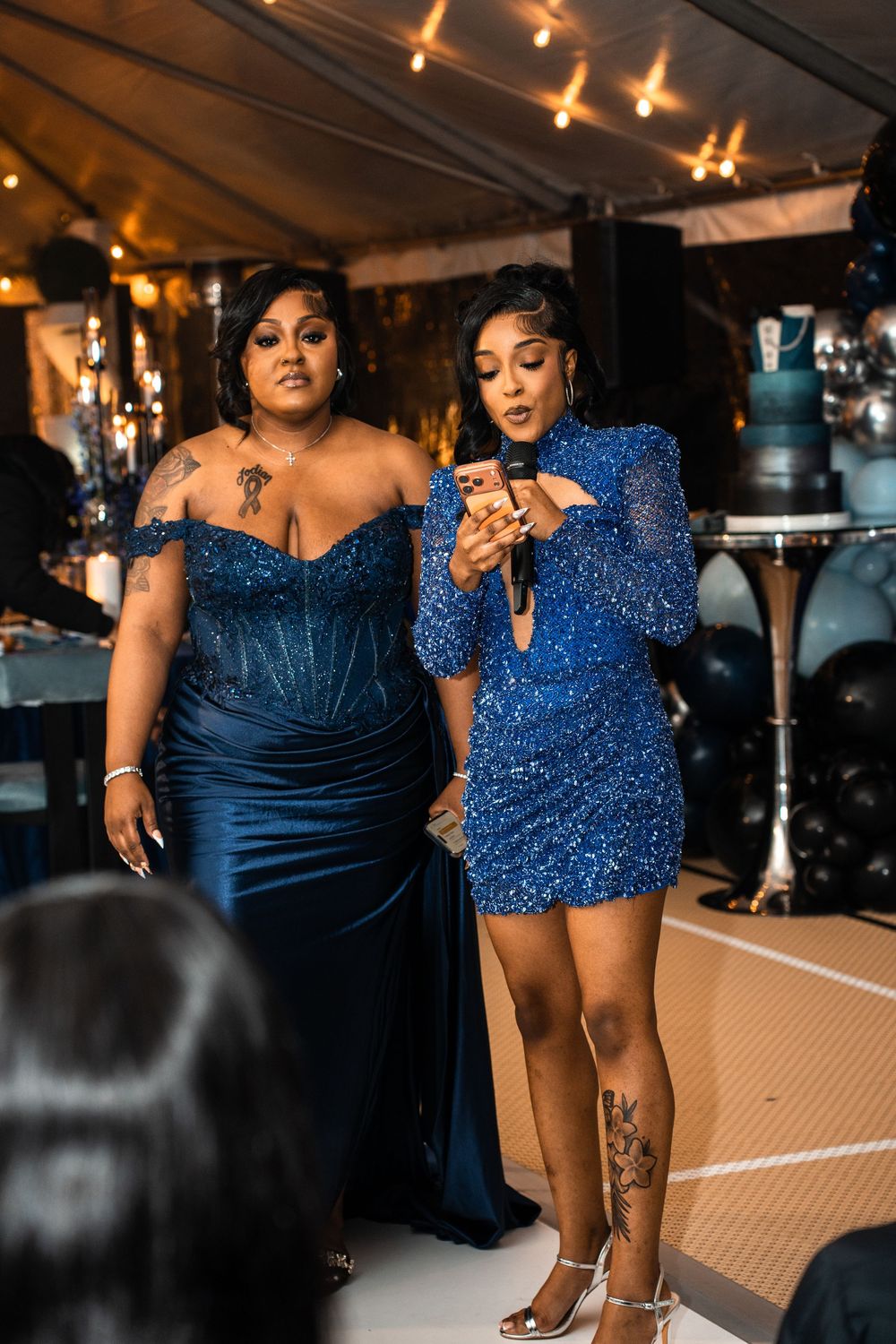 Two women in blue sequin dresses at an elegant event, one holding a microphone under a decorated tent.