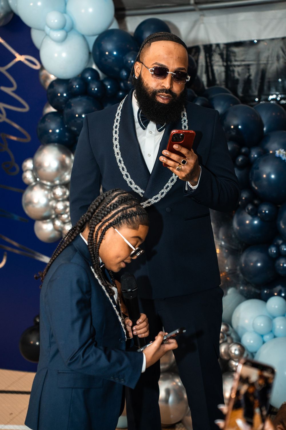 Man in navy suit with chain necklace uses phone beside girl with braids at balloon-decorated event.