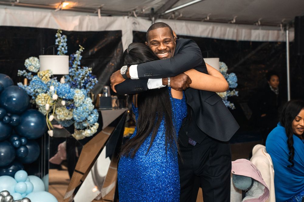 Two people sharing a warm hug at a blue-themed celebration event with balloon decorations and floral arrangements.