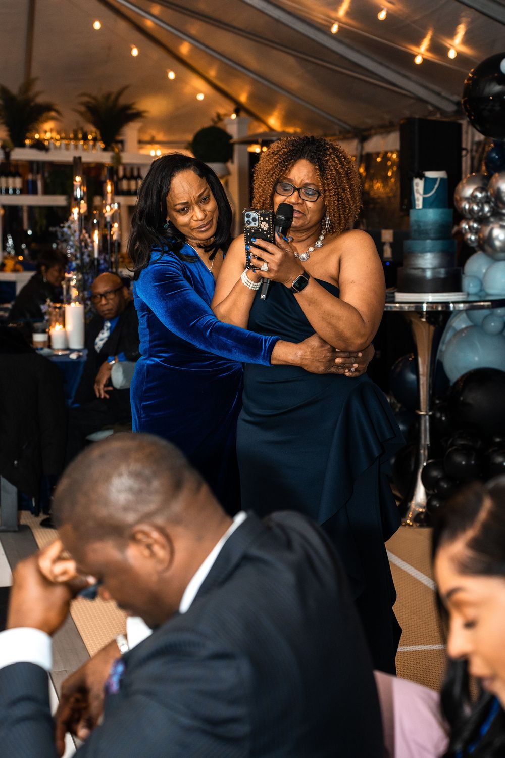 Two women in elegant blue gowns take a selfie at a formal evening event under a decorated tent.