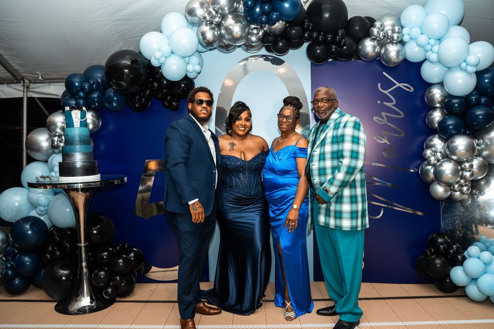 Four people posing together at a blue-themed party with balloon arch and decorated cake.