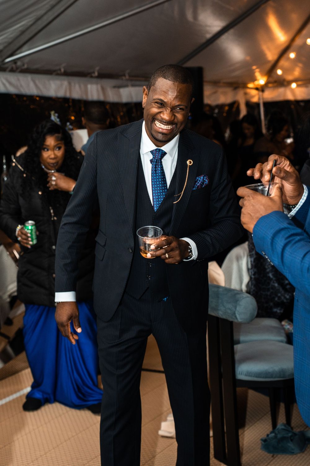 Smiling man in navy suit holding a drink at an elegant event under a white tent.