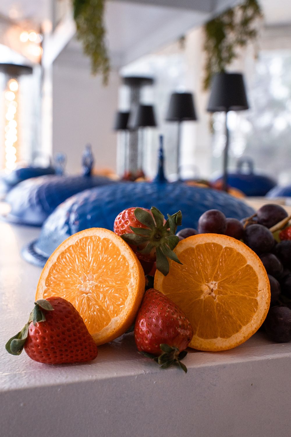 Sliced oranges, strawberries, and grapes on a white surface with blue decorative dishes and lamps in the background.