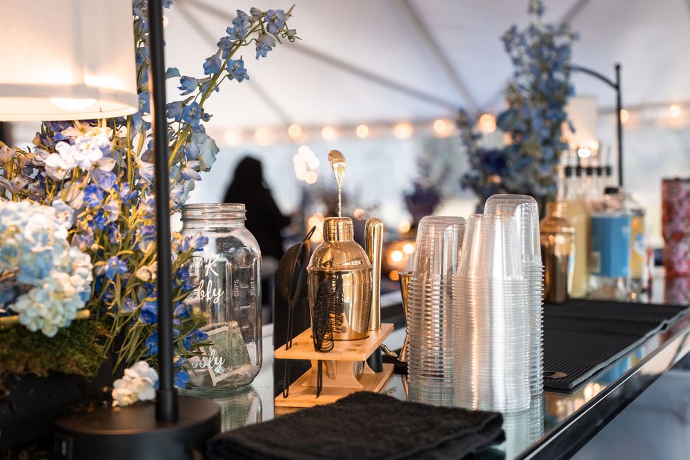 Elegant outdoor bar setup with blue floral arrangements, glass jars, and stacked cups under a white tent.