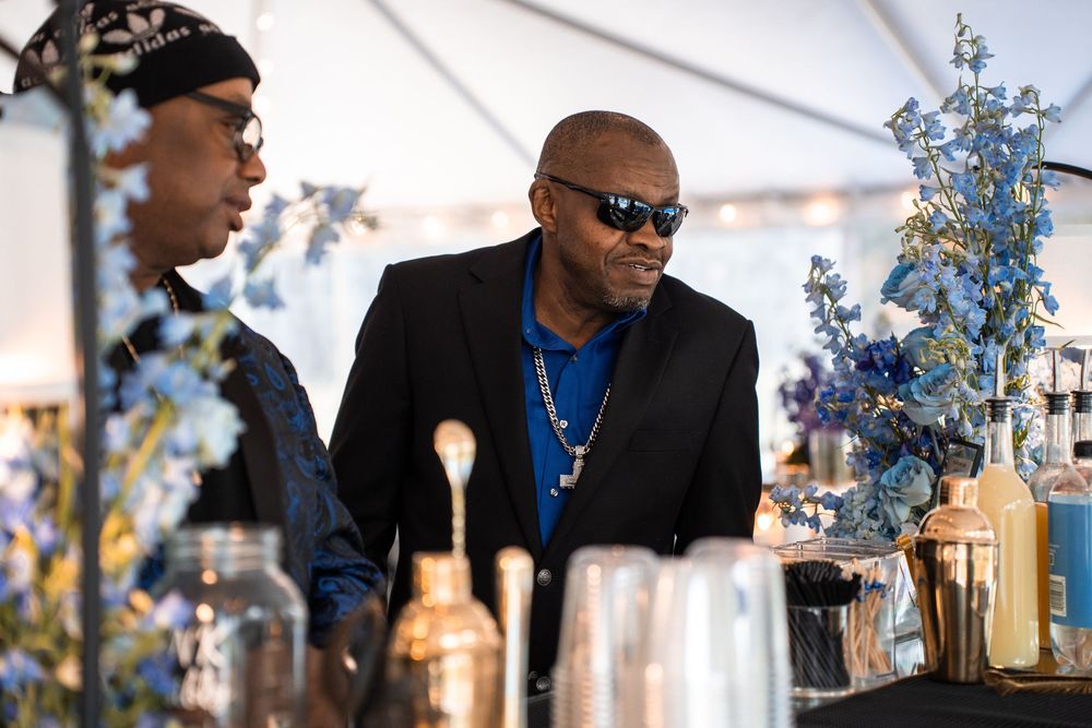 Two men at an elegant blue floral event bar under a white tent, one wearing sunglasses and a black blazer.