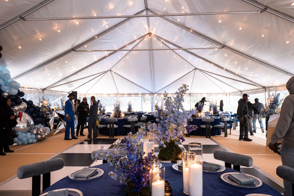 Elegant tented event with navy blue tablecloths, blue floral centerpieces, candles, and string lights overhead.
