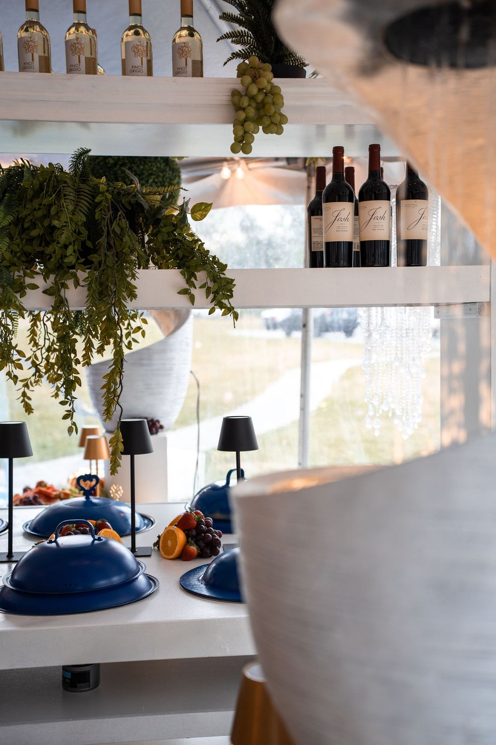 Elegant restaurant shelf display with red wine bottles, hanging greenery, and blue covered dishes on a white counter.
