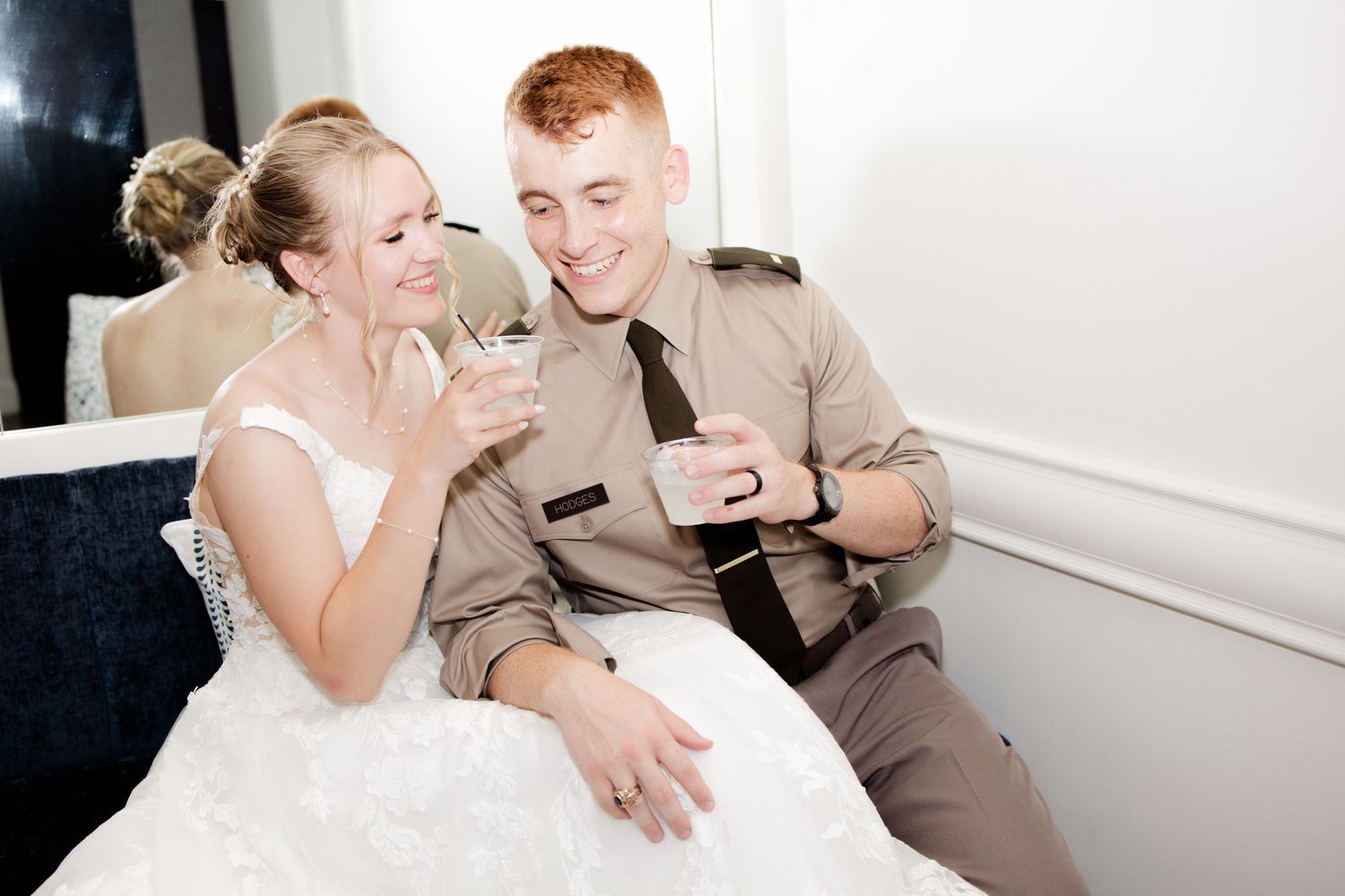 Bride in white gown and groom in military uniform share a joyful moment with drinks at their wedding reception.